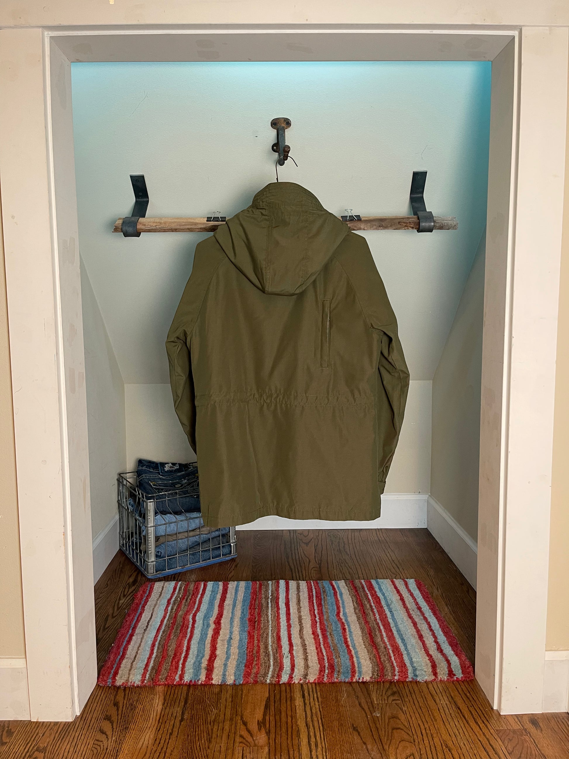 Back view green jacket hanging on a hook in a room with a striped rug on the floor.